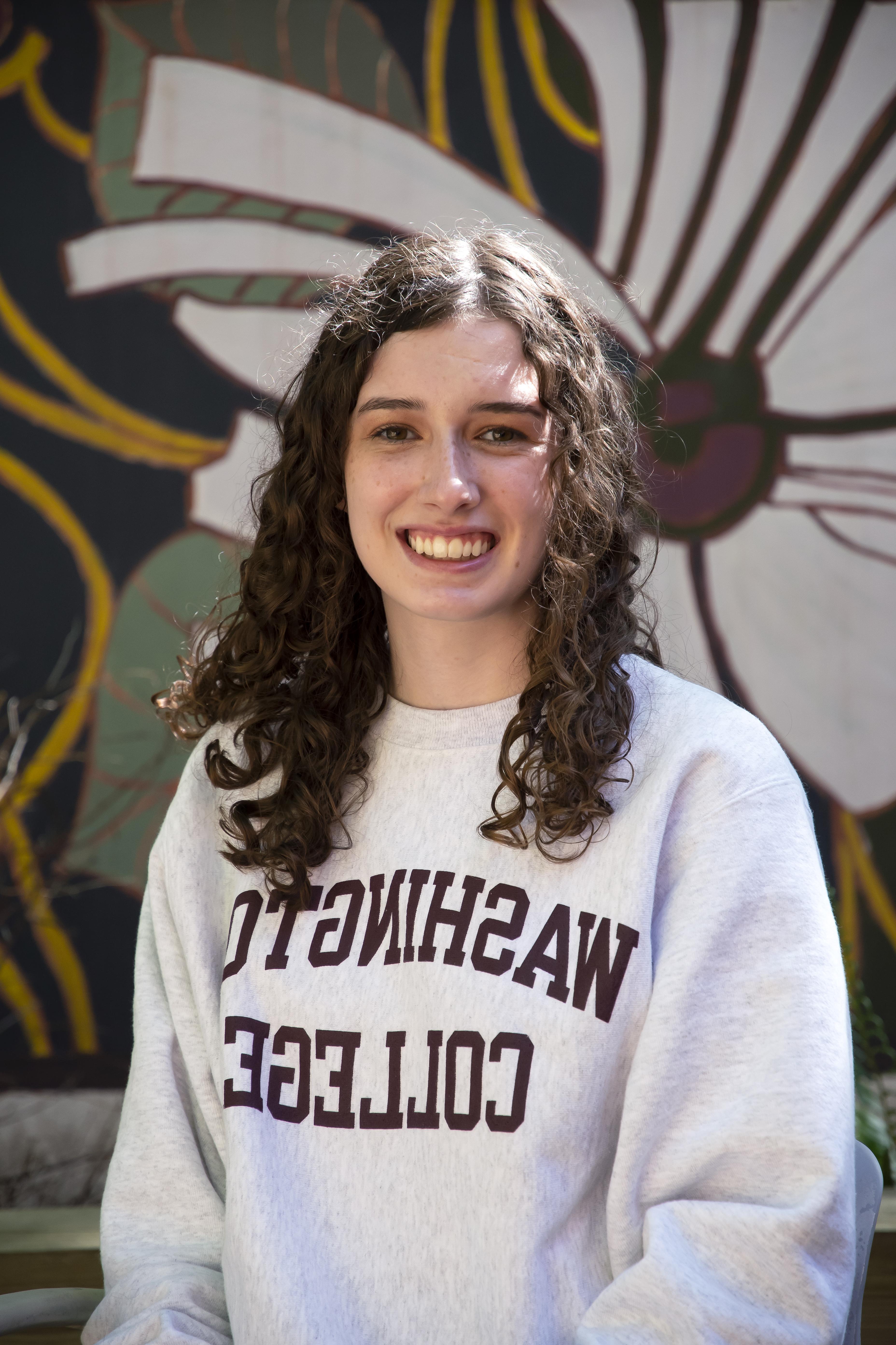 Emma, smiling and wearing a white Washington College sweatshirt with a painted mural behind her. 